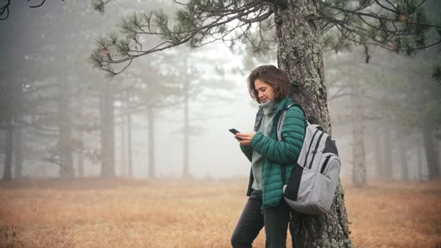 在公园里，一名年轻女子靠在一棵树上，用手机输入信息视频素材