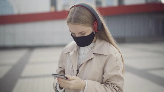 年轻的白人女性戴着口罩戴着耳机听音乐，用智能手机上网。冠状病毒大流行期间的现代城市生活方式。第二波的概念视频素材