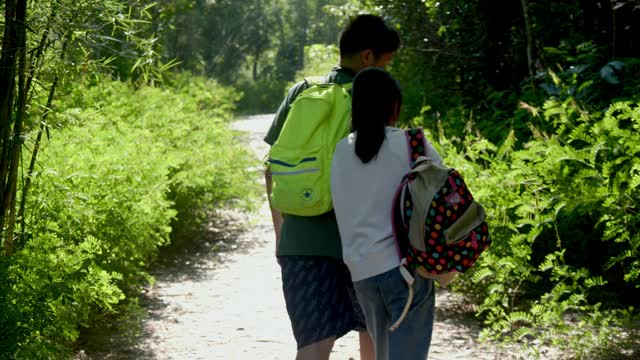 快乐亚洲男孩和他的妹妹背着背包一起在阳光明媚的日子里走在森林里，生活理念。视频素材