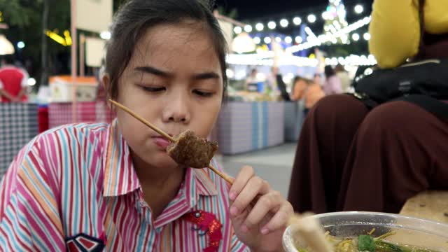 4K，亚洲女孩吃泰国街头食物叫猪肉烤木托盘在夜市的人走视频素材