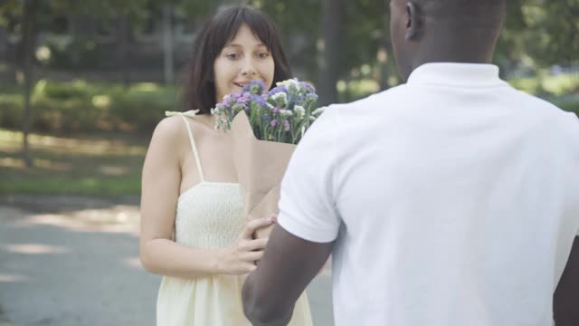 一个快乐的白人年轻女子在户外接受来自非裔美国人的花束。美丽的女朋友与爱的男友在阳光明媚的夏天约会。浪漫和爱情。视频素材