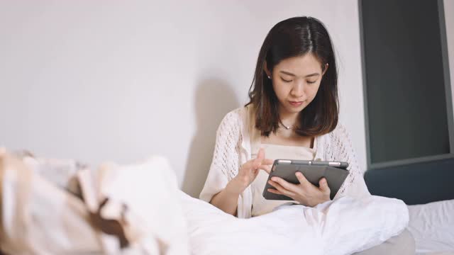 年轻的亚洲女人在网上使用智能手机平板电脑，她躺在家里的床上，亚洲女孩，放松和度假的概念。视频素材