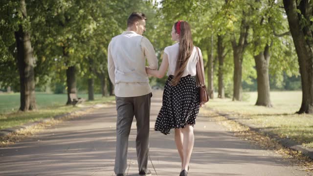 年轻夫妇在阳光明媚的夏天公园约会的广泛镜头。后视图苗条的年轻女子和优雅的男子散步和交谈。快乐的女友和男友边走边聊天视频素材