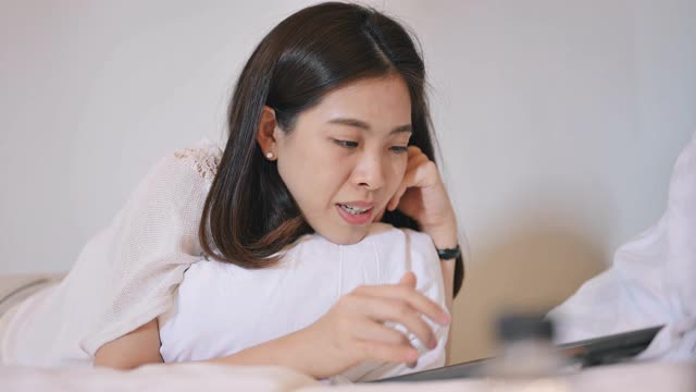 美丽微笑的亚洲职业妇女的肖像躺在她的床上用平板电脑智能手机和她的朋友打视频电话，在晚上和亲戚朋友聊天，买衣服的电子商务视频素材