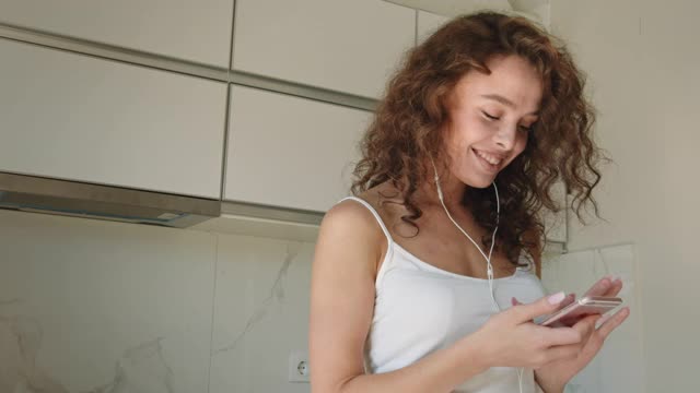 一位年轻女子戴着耳机听音乐，笑着。她在厨房里。她随着音乐跳舞。4 k。视频素材