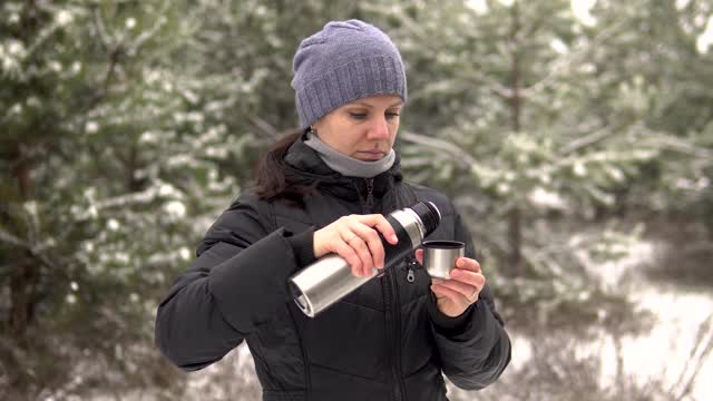 在一个下雪天，一个年轻女子站在森林或公园里。她从金属保温瓶里倒了一杯热饮。视频素材