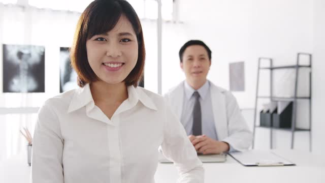 自信的亚洲男医生在白色医疗制服和年轻的病人女孩看着相机和微笑，而医疗咨询的桌子在健康诊所或医院。视频素材