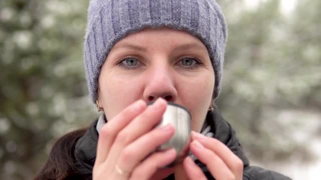 在一个下雪天，一个年轻女子站在森林或公园里。她正在喝热茶。她微笑,笑了。视频素材