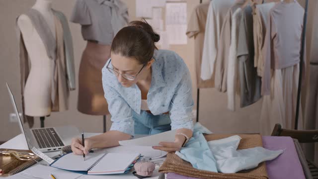 年轻女子时装设计师专注地工作，弯在桌子上，使衣服的草图和试织物上的人体模型视频素材