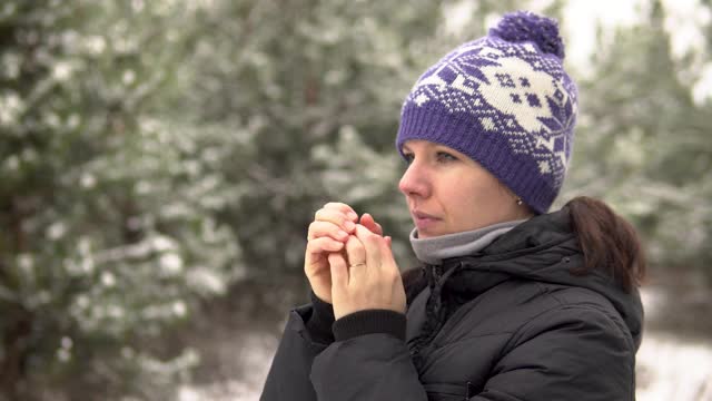 在一个下雪天，一个女孩站在森林或公园里，用她的呼吸摩擦和温暖她的手。视频素材