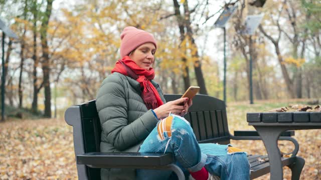 一个年轻的女人坐在秋天公园的长椅上用她的智能手机视频素材