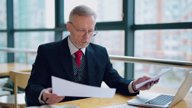 身着正装的资深商人肖像。一个成熟的男人坐在桌子前的笔记本电脑前，在靠近窗户的办公室里处理文件。视频素材