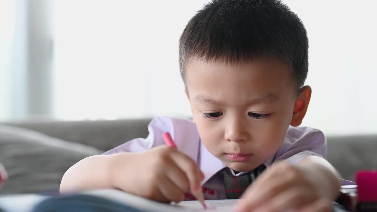 亚洲男孩做家庭作业的意图。一个男孩拿着铅笔写，一个男孩在白纸上画在桌子上，小学和家庭教育，远程在线教育概念。视频下载