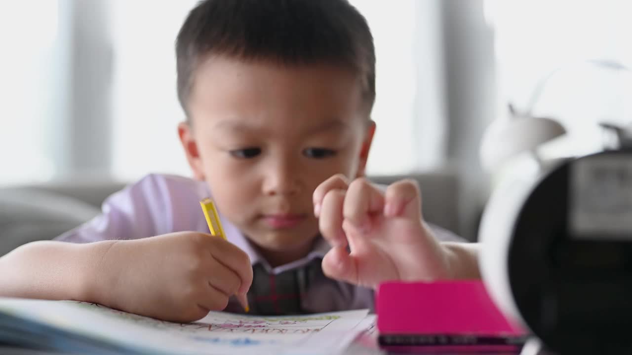 亚洲男孩做家庭作业的意图。一个男孩拿着铅笔写，一个男孩在白纸上画在桌子上，小学和家庭教育，远程在线教育概念。视频下载