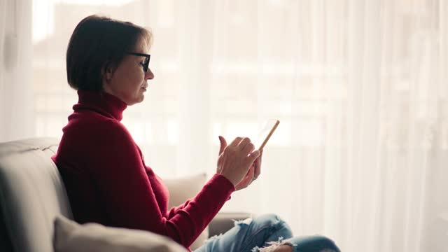 放松的年轻女子戴着眼镜，坐在沙发上用她的智能手机。视频素材