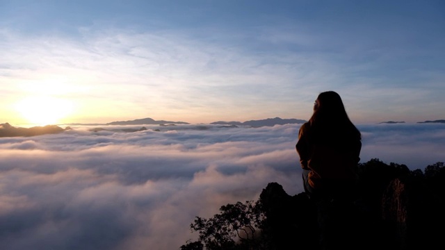 一个女旅行者坐在山顶上看日出和雾海的慢镜头视频素材