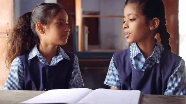 两名穿着蓝色格子制服的女同学坐在桌子上，彼此挨着，一边讨论一边举起手来掩盖出席情况视频素材