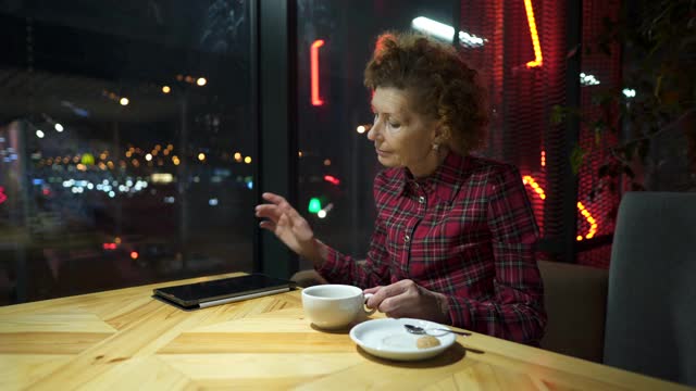 成熟的白人女人喝着咖啡，看着平板电脑，上网。晚上坐在城市咖啡馆里。老年女性在咖啡店使用科技，坐在窗口与城市景观视频素材