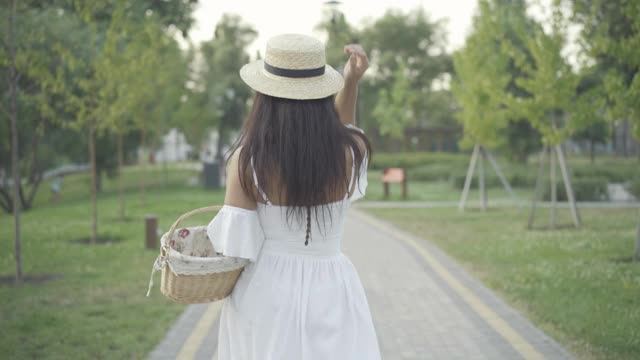 美丽苗条的中东妇女穿着白色连衣裙和草帽纺纱漫步在阳光明媚的夏日公园小巷。肖像积极快乐的女士走在户外野餐篮子。视频素材