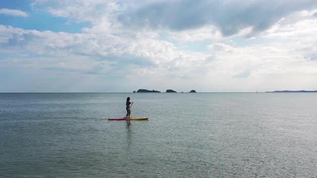 在夏日阳光明媚的日子里，自信亚洲女人站在冲浪板上，用4K无人机鸟瞰美丽的大海和天空。女性在假期喜欢水上娱乐运动视频素材