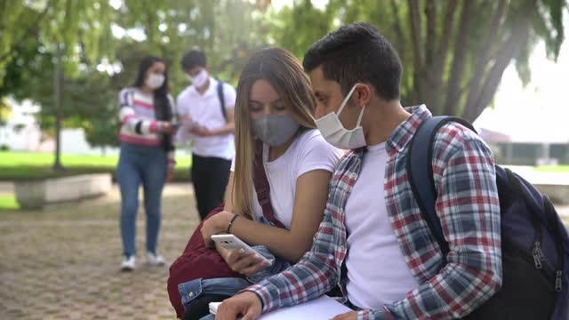 一对学生情侣在大学校园里一边聊天一边看智能手机，所有人都戴着防护口罩视频素材