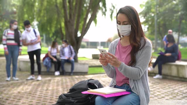 一名年轻女学生在校园里用智能手机发短信，看上去很开心，周围的学生都戴着防护口罩视频素材