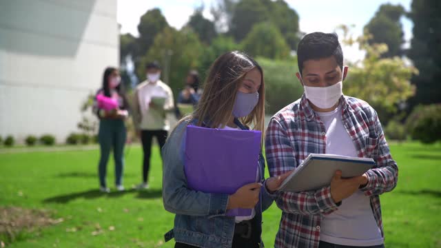 一对快乐的年轻情侣在大学校园里一起检查笔记本，当偶然的学生经过时，他们戴着防护口罩视频素材