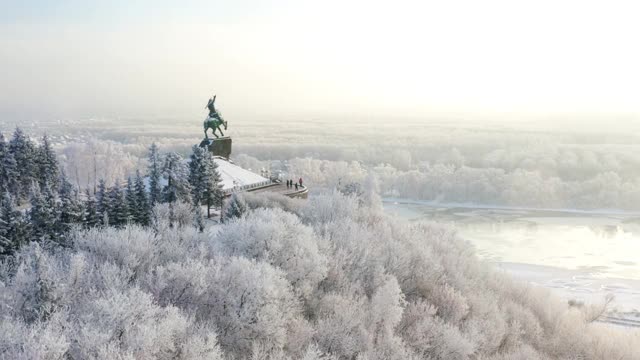 萨拉瓦特·尤拉耶夫纪念碑坐落在一座山上，周围是积雪覆盖的树木，背景是一条河流。冬季堤岸的美丽景色开始了。无人驾驶飞机。视频素材