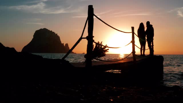 一对年轻夫妇在一个乡村码头上接吻，凝视着夕阳，背景上写着“Es Vedrà”。伊比沙岛- 4 k视频素材