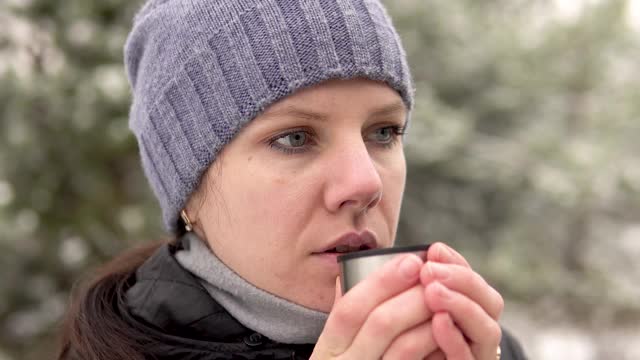 在一个下雪天，一个年轻女子站在森林或公园里。她正在喝热饮。视频素材