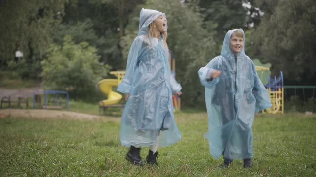 欢乐的孩子们跳跃着，用嘴接住雨滴。快乐无忧无虑的白人男孩和女孩的肖像在雨天的夏天户外玩的雨衣视频素材