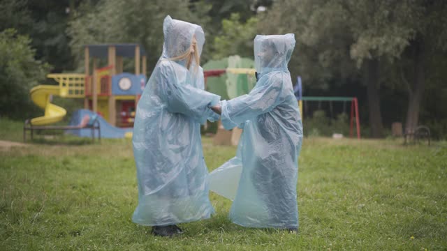 欢快的白种人孩子在户外旋转跳跃。快乐的女孩和男孩穿着雨衣在绿色的草地上玩耍在夏天的一天。欢乐和快乐的童年理念视频素材