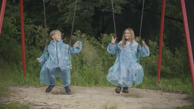 放松的孩子们穿着雨衣在户外荡秋千的广角镜头。正面微笑的白人男孩和女孩享受雨天的夏天。友谊和休闲的概念视频素材