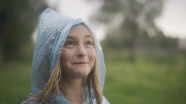 正面美丽的女孩在雨衣兜帽捕捉雨滴与舌头的特写。兴奋的白人女学生在户外享受夏天的雨视频素材