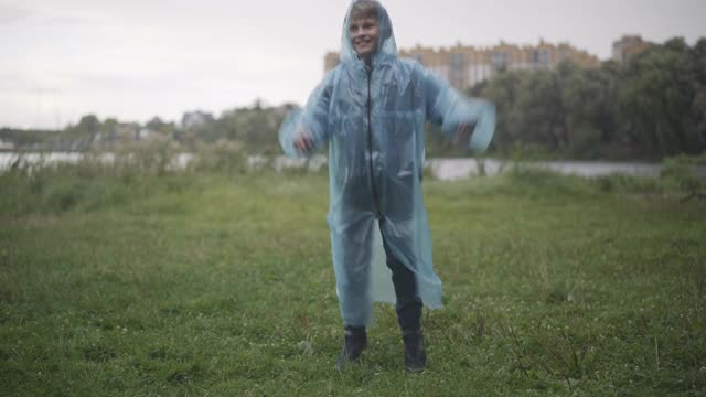 照相机接近快乐的白人男孩在雨衣有乐趣在雨天的夏天户外。活泼可爱的男生的肖像跳跃和旋转。快乐的童年概念视频素材