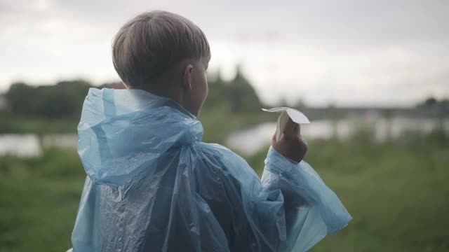 后视图可爱的正面男孩在雨衣和纸飞机沿着绿色的夏季草地跑。镜头跟随快乐的白人学童在阴天户外享受雨天视频素材