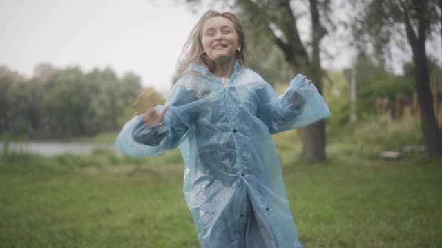 活泼的女学生在户外蹦蹦跳跳，穿上雨衣兜帽。快乐的白人小女孩享受雨天的夏天的肖像。童年的生活方式和休闲观念视频素材