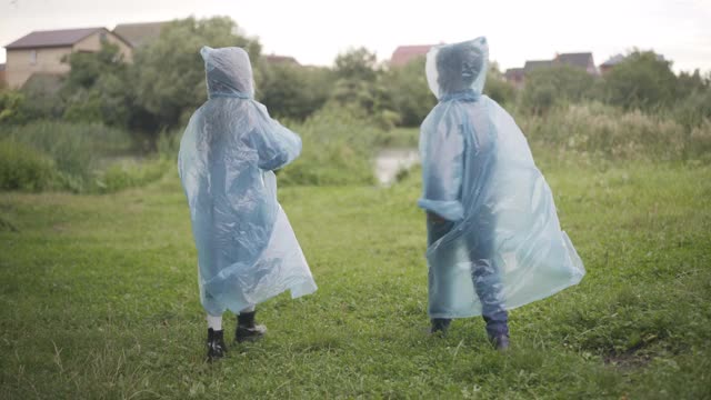 两个穿着雨衣的孩子在绿色的夏日草地上散步。愉快友好的白人朋友在户外散步和交谈的背影。童年和友谊视频素材