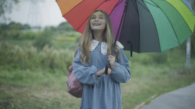 迷人开朗的女学生站在户外，拿着五颜六色的雨伞微笑着。正面的白人女孩在户外享受雨天的肖像。童年与幸福观视频素材