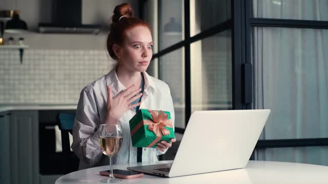 快乐的年轻女子打开礼盒与礼物，同时有视频电话上的笔记本电脑。视频素材