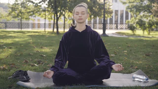 广角镜头冷静自信的白人妇女坐在荷花姿势的运动垫子上。肖像年轻的女性瑜伽修行者在户外的阳光训练。运动与宁静的概念。视频素材