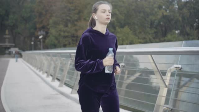 疲惫的白人女运动员在雾蒙蒙的天气里拿着水瓶跑步的肖像。精疲力竭的美女停止慢跑并叹息。锻炼和疲惫。视频素材