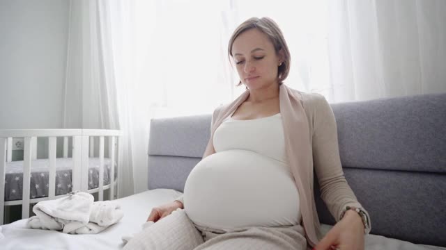 年轻孕妇穿着婴儿鞋坐在家里的沙发上。孕妇正在为分娩做准备。视频素材