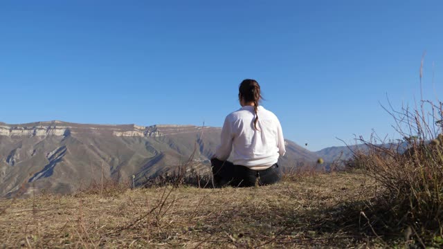 深褐色的女人穿着一件白色的衬衫坐在地上，在山上的晴朗的天空下视频素材