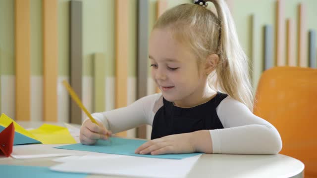 可爱的金发白种女孩在幼儿园或学校的桌子上用铅笔画画。肖像快乐的小学生画画和微笑。童年和教育。视频素材