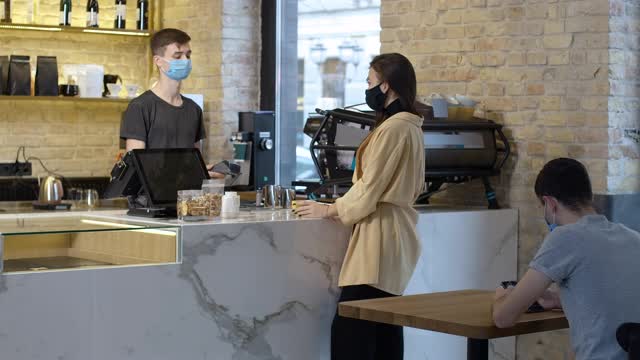 年轻的白人褐发女人戴着冠状病毒面具在咖啡馆买外卖咖啡然后离开。咖啡师在室内为客户服务。新常态冠状病毒生活方式视频素材