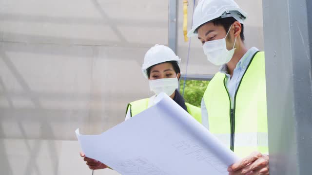 亚洲工人在建筑现场佩戴防护口罩。男和女工程师拿着图纸在施工方讨论项目。视频素材