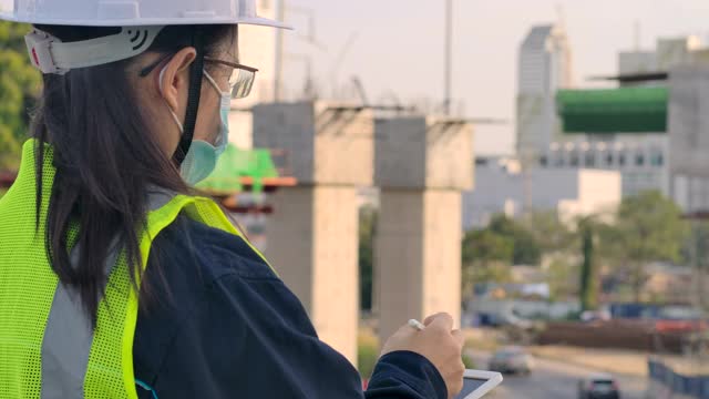 亚洲女性土木工程师专家或建筑建筑师戴着安全安全帽在房地产建筑项目上工作，在城市建设现场检查和记录建设进度碑。视频素材