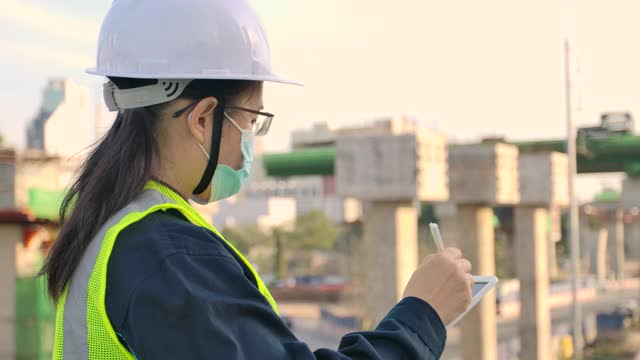 房地产建设业务。亚洲女性土木工程师或建筑建筑师戴安全帽在城市建筑工地检查和记录施工进度视频素材