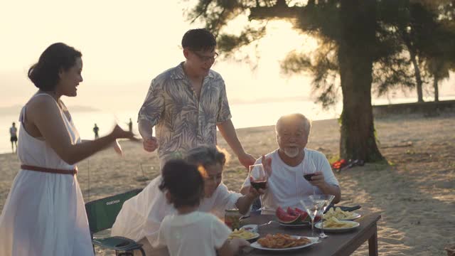 快乐的亚洲家庭在海滩度假。一群多代同堂的家庭在夏日日落的海滩上享受晚餐聚会和跳舞。小女孩与父母和祖父母一起放松和快乐。视频素材